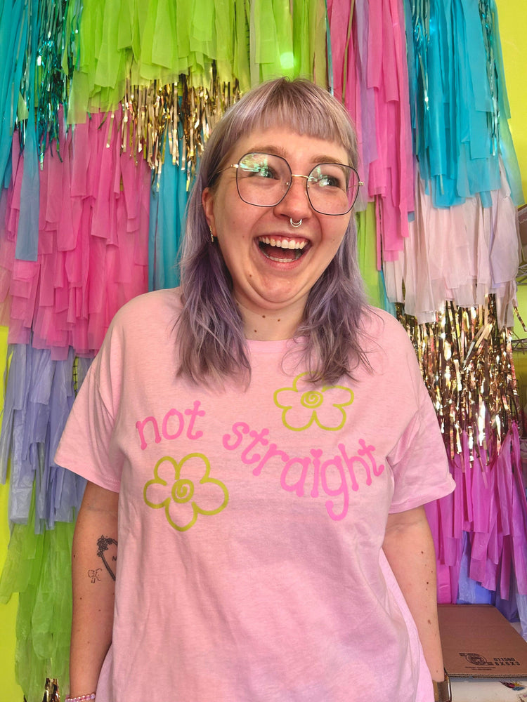 Person wearing a pink t-shirt with 'not straight' text and flowers, standing in front of colorful streamers.
