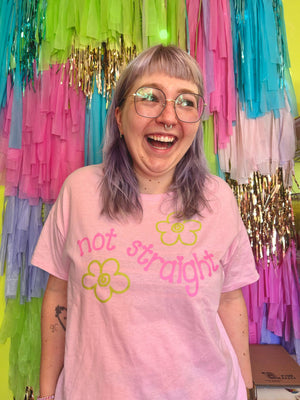Person wearing a pink t-shirt with 'not straight' text and flowers, standing in front of colorful streamers.