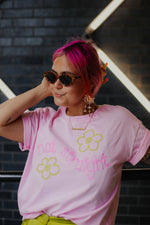 Person wearing a pink t-shirt with floral designs and sunglasses, standing against a dark brick wall.