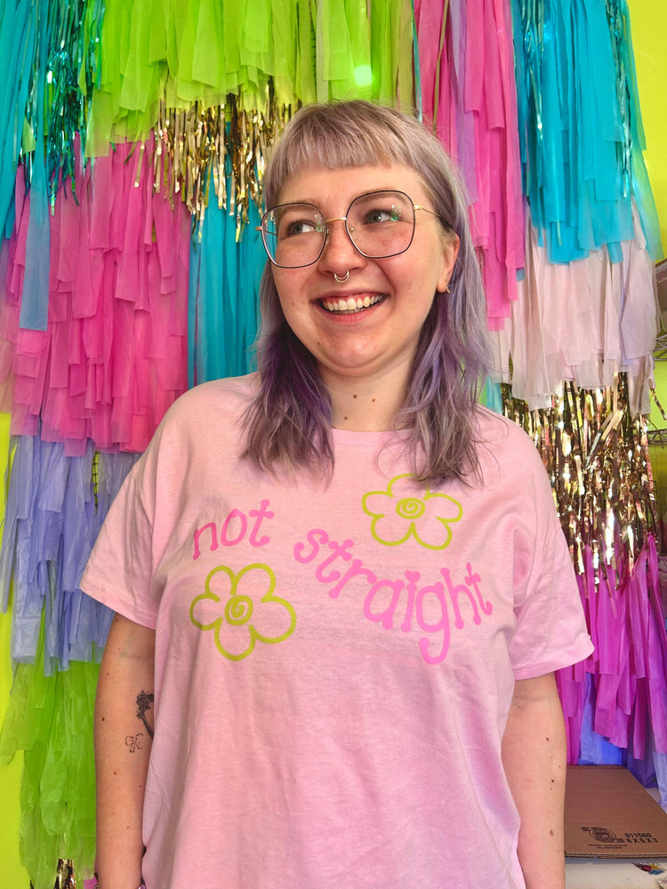 Person wearing a pink t-shirt with 'not straight' and flower graphics in front of colorful streamers.