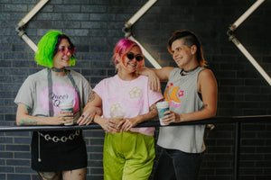 Three people with colorful hairstyles and clothing standing together against a dark brick wall.