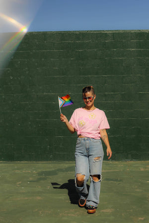 Person holding a rainbow flag against a green wall with a rainbow light effect.
